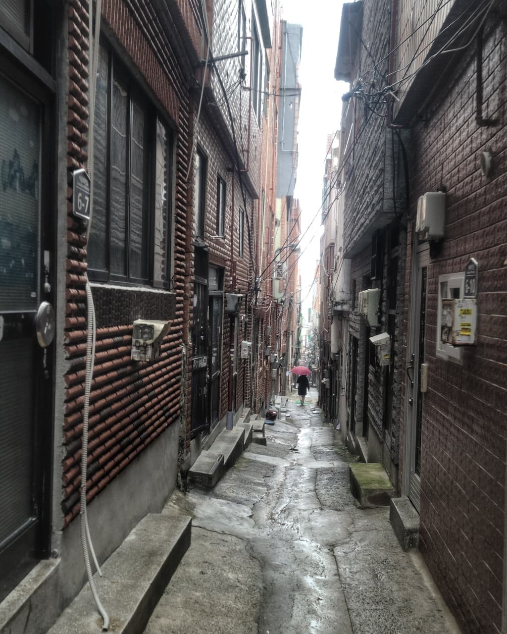 비 온 뒤 벽돌집 사이로 난 좁은 골목. 멀리 우산 쓴 사람이 걸어간다.